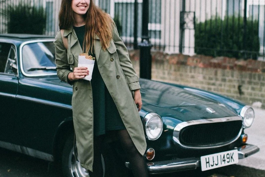 Woman in a gray trench coat resting on a car