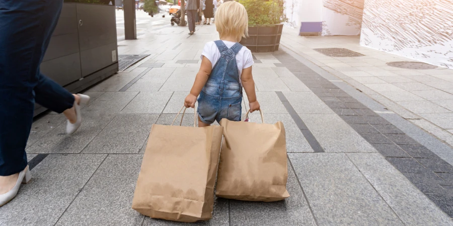 Little cute blonde kid filled a lot of shopping bags in the street after the Black Friday sales. View from the back. The toddler is happy, she is a shopaholic.
