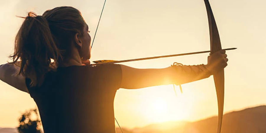 Woman pointing a bow and arrow in direction of sunset