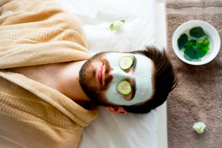 Um homem de roupão deitado usando uma máscara facial e pepinos nos olhos