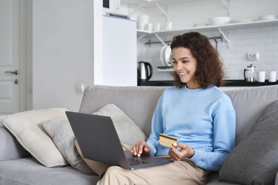 A woman sits on the sofa in her apartment looking at a computer and holding her credit card
