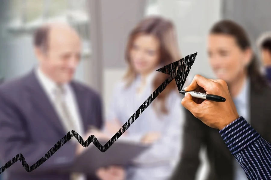 A person drawing an up arrow on a transparent board with office workers blurred in the background