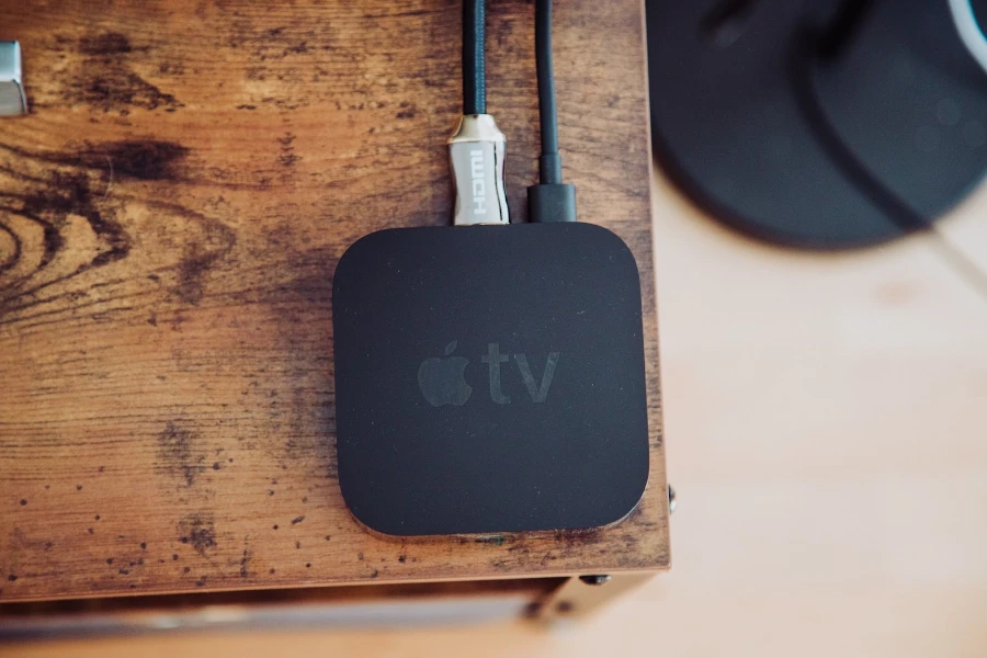 Black Apple TV sitting on a wooden table with two cords plugged into it
